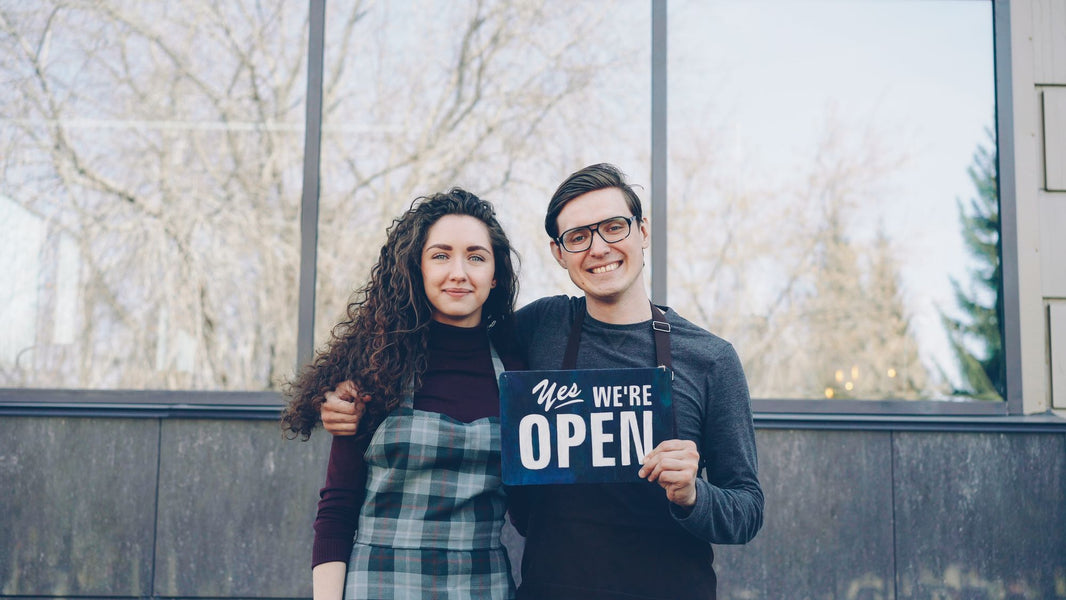 Zwei Ladeninhaber stehen lachend mit einem Open-Schild vor ihrem Geschäft